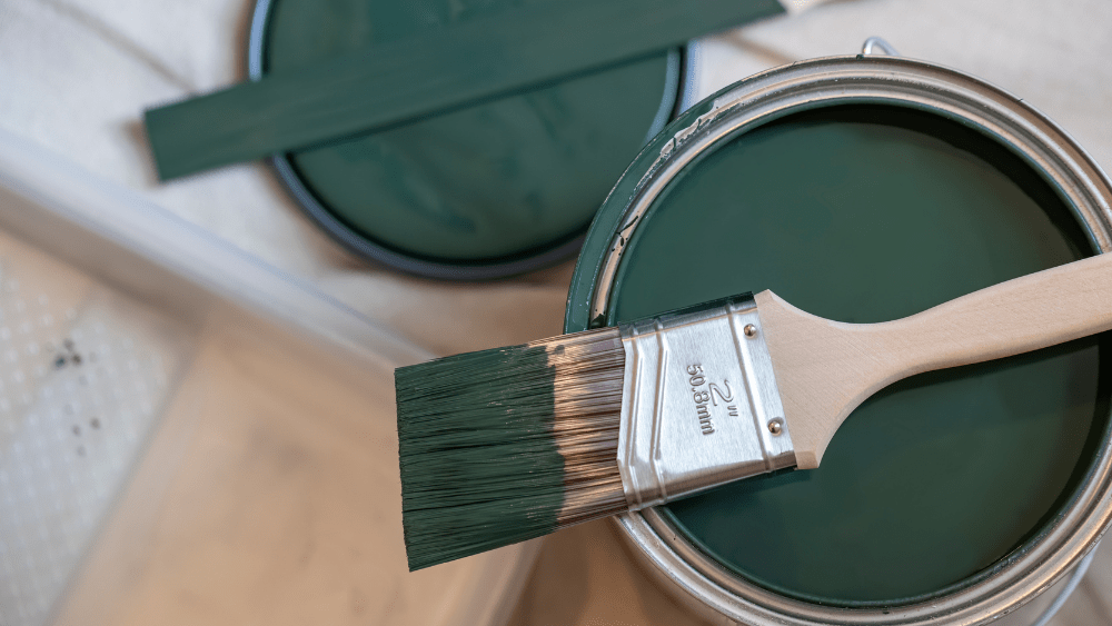 Open paint can with dark green paint and a paint brush laying across the top.